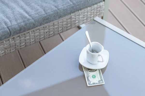 American one dollar bill and empty cup of coffee on a glass table of outdoor cafe. Payment, tip.