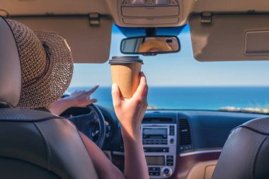 Hasır şapkalı sürücü kadın elinde kağıt bir fincan kahve yle araba ile seyahat ediyor. Ön cam ve deniz arka koltuktan görünüm