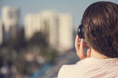 Tatil kasabasının arka planına karşı yaz sabahı kulaklıkla müzik dinleyen pembe gömlekli kadının arka görünümü