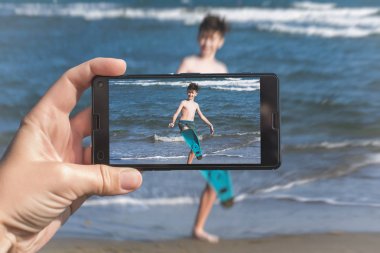 Female taking a picture of a boy on the beach on the phone. Happy teen boy having fun on the sand n the beach.Travel and family concept clipart