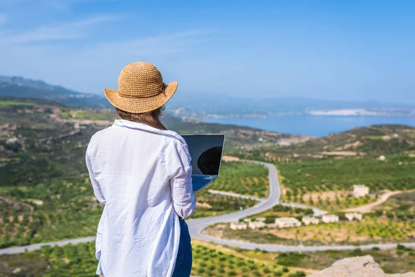 Woman freelancer in tourist clothes and straw hat with a laptop on top of a mountain. Concept