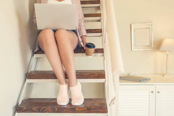 Girl student in shorts sits on the steps with a laptop and paper cup of coffee in a cafe in the home style