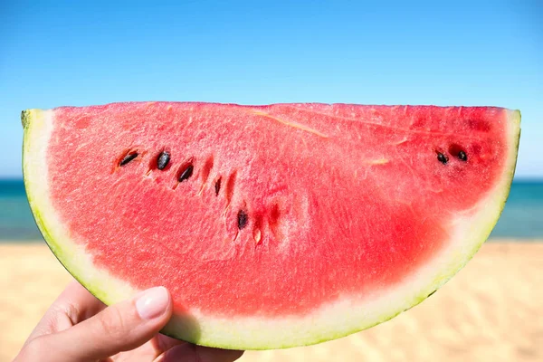 Ripe piece of watermelon in female hands on the background of the beach on a hot summer day. Concept