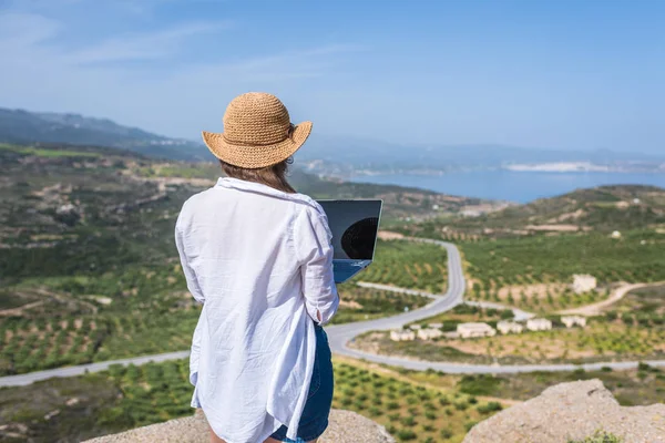 Woman freelancer in tourist clothes and straw hat with a laptop on top of a mountain. Concept