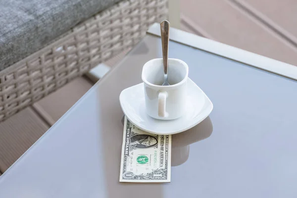 American one dollar bill and empty cup of coffee on a glass table of outdoor cafe. Payment, tip.