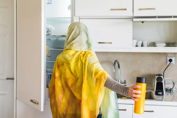Back view on a muslim woman in a scarf taking out juice from the kitchen fridge. Breakfast preparation