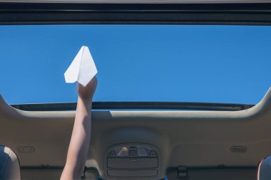 A girl is launching a paper airplane from open hatch of the car during summer vacation. Travel and freedom concept clipart