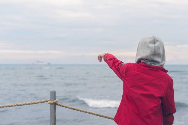 Kırmızı ceketli ve kulaklıklı bir çocuk fırtınalı yağmurlu havalarda deniz dalgalarına bakıyor.