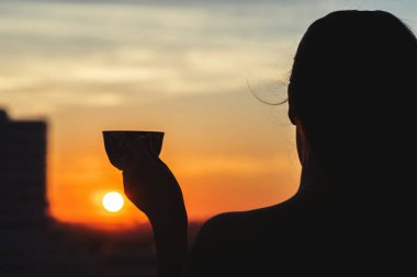 Silhouette of girl with a cup of tea at sunset. View on the town from above  clipart