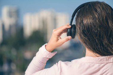Tatil kasabasının arka planına karşı yaz sabahı kulaklıkla müzik dinleyen pembe gömlekli kadının arka görünümü