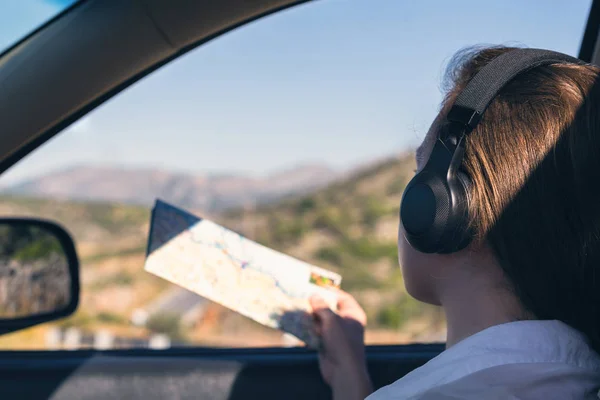 Young woman traveling by car listen music in the headphones and looks  on the map in the open window of the auto