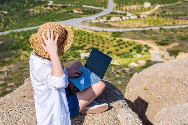 Woman freelancer in tourist clothes and straw hat with a laptop on top of a mountain. Concept