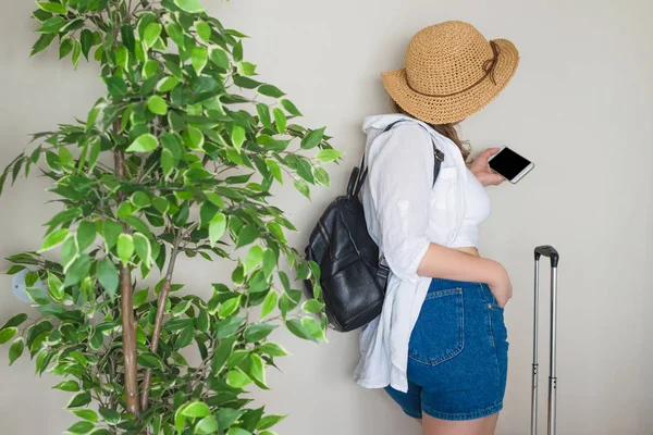 A lonely woman traveler standing in a waiting area of airport with suitcase and mobile phone near the wall and big green tree