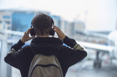 Bir sırt çantası ile kulaklık bir çocuk havaalanında pencerenin yanında duruyor ve uçağa bakar. Gidiş salonu