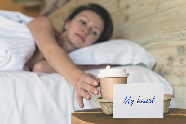 A paper cup of coffee and sticker with phrase "my heart" standing on the table in the bedroom as a surprise for a sleeping girl. 