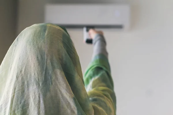 穆斯林妇女拿着一条围巾 从房间内的空调遥控器 — 图库照片