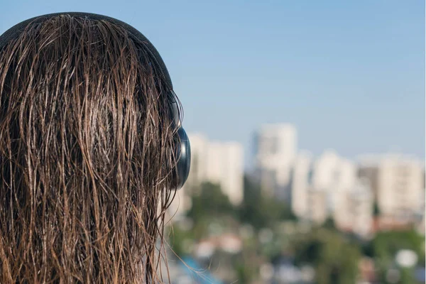 Back view of woman with wet hair in a headphones on the balcony of her apartment against the backdrop of the resort town and the sea.