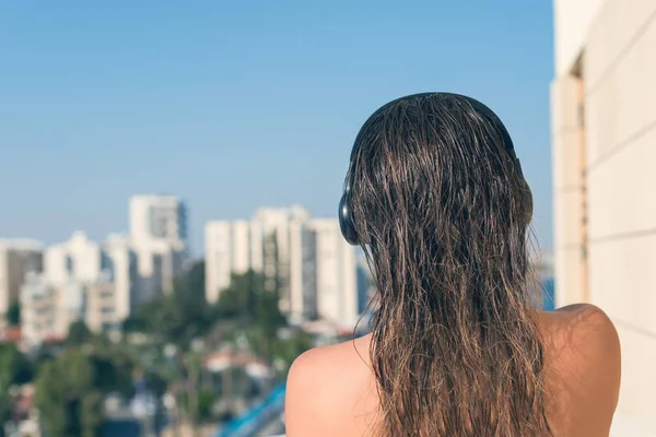 赤裸裸的美丽女人与湿头发在耳机在她的公寓的阳台上 在度假小镇和大海的背景 — 图库照片