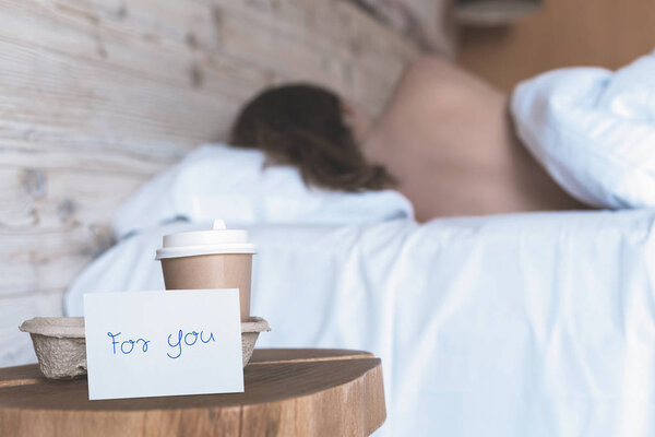 A paper cup of coffee and sticker with phrase "for you" standing on the table in the bedroom as a surprise for a sleeping girl. 