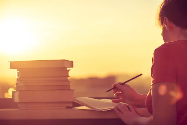 Female student with a lot of books preparing for university exam late evening on the sunset town background. Education concept