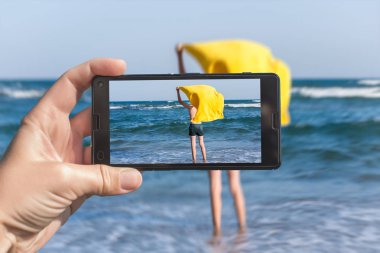 Kadın, plajda bir çocuğun resmini çekerken telefonda. Kumlu n plajda eğlenmek mutlu genç çocuk. Seyahat ve aile konsepti