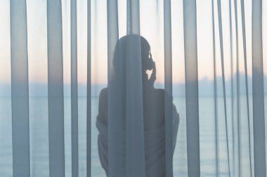 Kulaklık çıplak güzel kadın ve gün doğumunda cam balkonda şeffaf perde arkasında dans. Deniz manzarası.