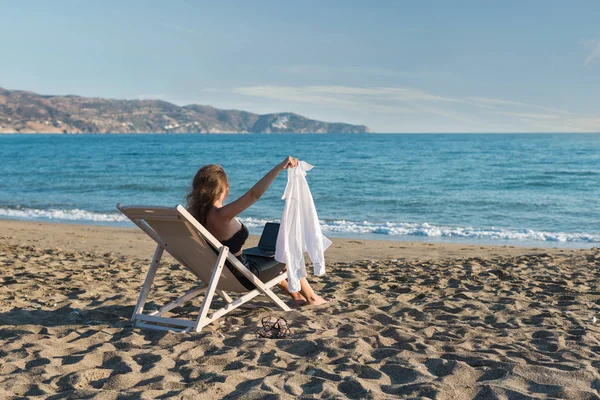 A young woman in office clothes with a laptop on the beach on a background of the sea in a summer sunny day. Concept