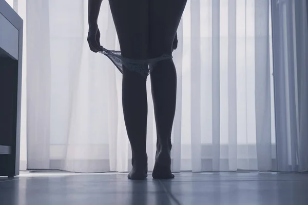 カーテンを背景に部屋で彼女の白いパンティーを脱ぐセクシーな脚を持つ美しい女性のシルエットの下のビュー — ストック写真