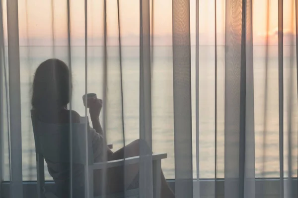 在日出时分 在玻璃阳台上透明窗帘后面 穿着毯子的裸女裹在椅子上 喝着咖啡 — 图库照片