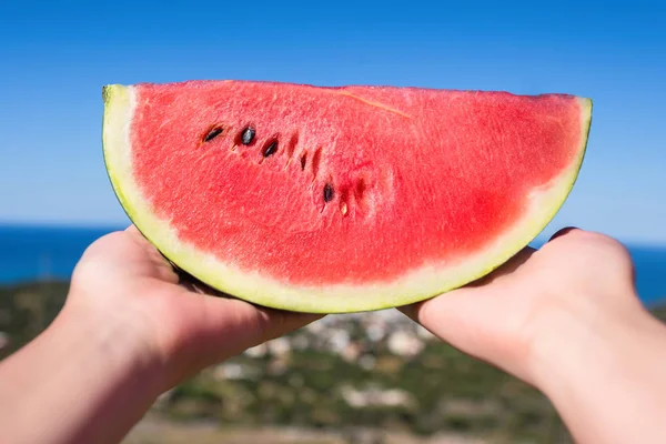 Ripe piece of watermelon in female hands on the background of the sea on a hot summer day. Concept