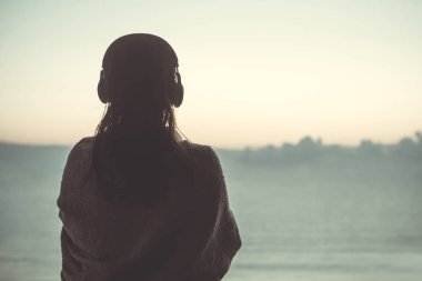 Çıplak kadın silueti ıslak saçlı bir battaniyeye bardı yüzdükten sonra. Denizkarşısı sahilde duran kulaklıklı kadın.