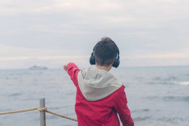 Kırmızı ceketli ve kulaklıklı bir çocuk fırtınalı yağmurlu havalarda deniz dalgalarına bakıyor.