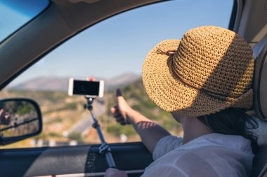 Blogger kız hasır şapka lı akıllı telefon ve selfie çubuğu kullanarak selfie fotoğrafı veya video çekiyor. Tatil ve turizm konsepti