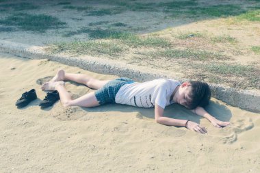 Kumsalda tişört lü ve şortlu bir çocuk yatıyor. 