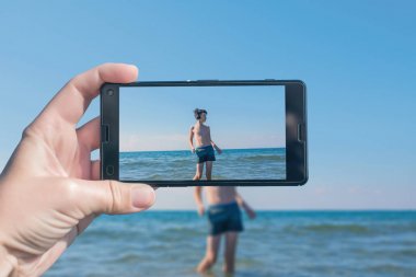 Kadın, plajda bir çocuğun resmini çekerken telefonda. Kumlu n plajda eğlenmek mutlu genç çocuk. Seyahat ve aile konsepti