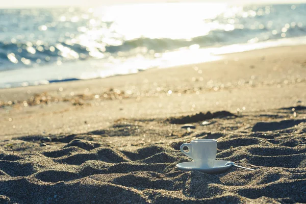 A cup of coffee in the sand at sunrise near the sea. Concept