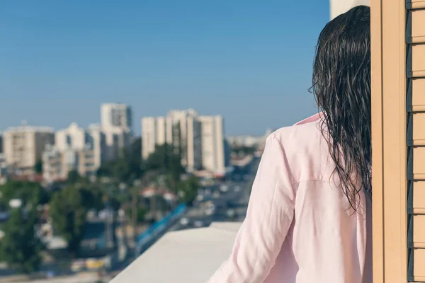 Back view of woman in a pink shirt with wet hair standing on the balcony at the summer morning against the background of resort town