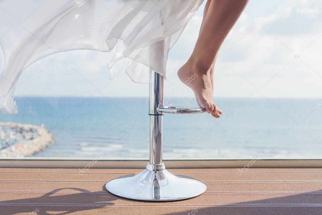 woman is sitting on a bar stool in a summer cafe on the sea and sky background. Bottom view on barefoot female legs