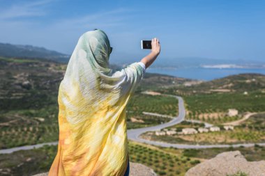 Renkli bir eşarp Müslüman kadın gezgin bir dağın tepesinde akıllı telefon üzerinde kendi portresini çekmek.