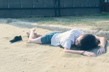 Kumsalda tişört lü ve şortlu bir çocuk yatıyor. 