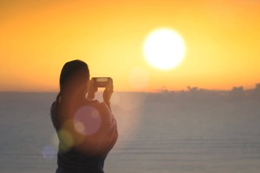 Yüzdükten sonra battaniyeye sarılmış ıslak saçlı kadının silueti. Gün batımı denizin karşısındaki sahilde duran cep telefonuyla fotoğraf çeken kadın.