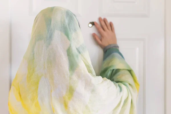 muslim woman in a colorful scarf looking on peephole door when somebody rings the doorbell