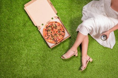 Bahçede çimenlerin üzerinde piknik yapmak. Beyaz yazlık elbiseli, bir kutuda pizza ve bir kadeh şarap la genç bir kadının havadan görünümü