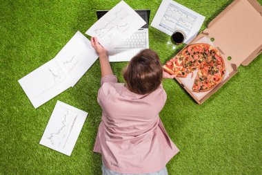 Gazeteye basılmış grafik ve grafiklerle dizüstü bilgisayar kullanarak çimüzerinde çalışan pizza ve bir kadeh şarapla öğle yemeği alan kadın.