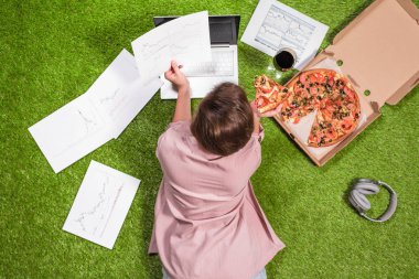 Gazeteye basılmış grafik ve grafiklerle dizüstü bilgisayar kullanarak çimüzerinde çalışan pizza ve bir kadeh şarapla öğle yemeği alan kadın.