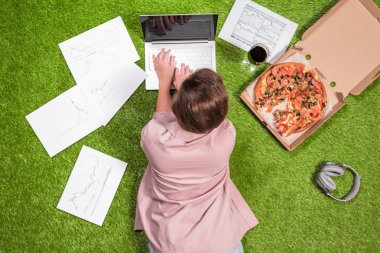 Gazeteye basılmış grafik ve grafiklerle dizüstü bilgisayar kullanarak çimüzerinde çalışan pizza ve bir kadeh şarapla öğle yemeği alan kadın.