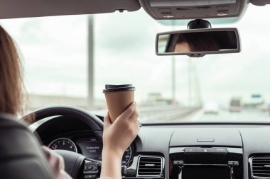 Kadın sabah otoyolda araba sürüyor ve elinde bir fincan kağıt kahve tutuyor. Arabanın arka koltuğundan bak. 