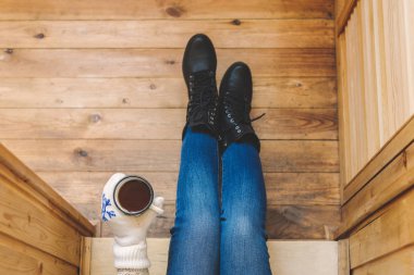 Ahşap bir evin terasında demir bir fincan kahve içen çizmeli bir kız. Kış konsepti