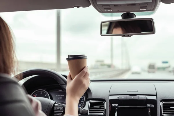 女性は高速道路を運転して午前中にコーヒーの紙コップを持っています 車の後部座席からの眺め — ストック写真