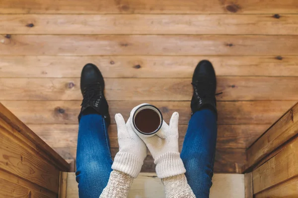 A girl in the boots with an iron mug of coffee on the terrace of a wooden house. Winter concept